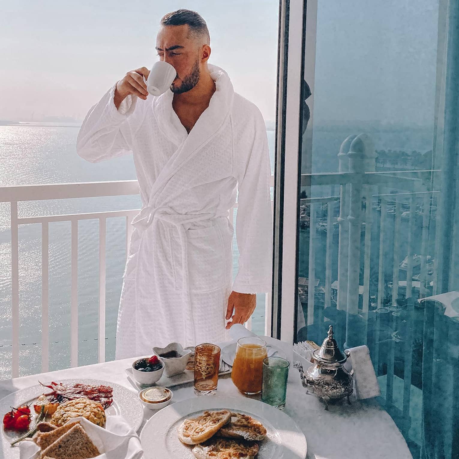 Man wearing white bath robe sips coffee in front of balcony breakfast table