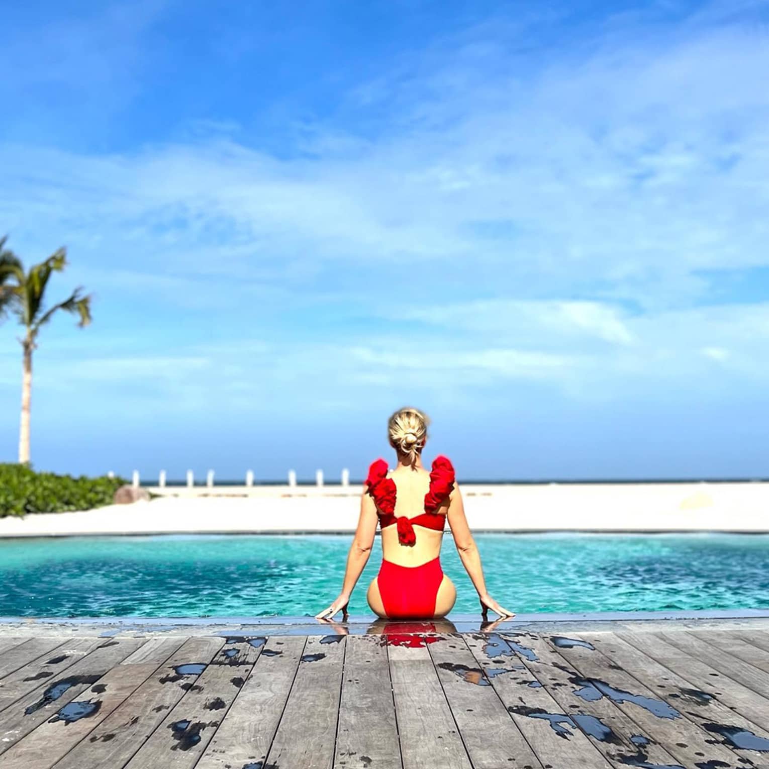 A person in a red swimsuit sits at the edge of a pool, gazing at the ocean under a clear blue sky, creating a serene and picturesque scene.