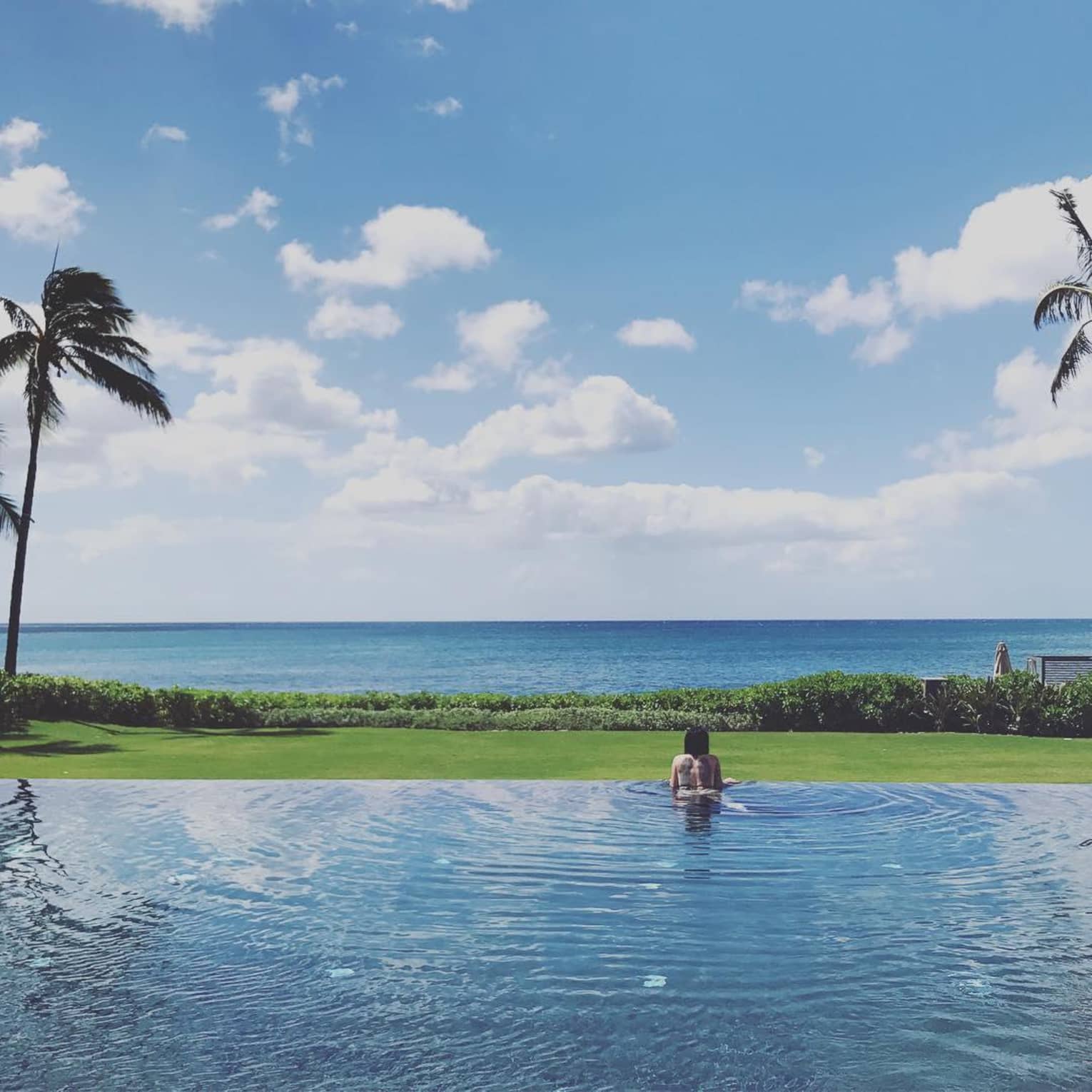 Woman wades in water at edge of swimming pool, looks out at green lawn, ocean