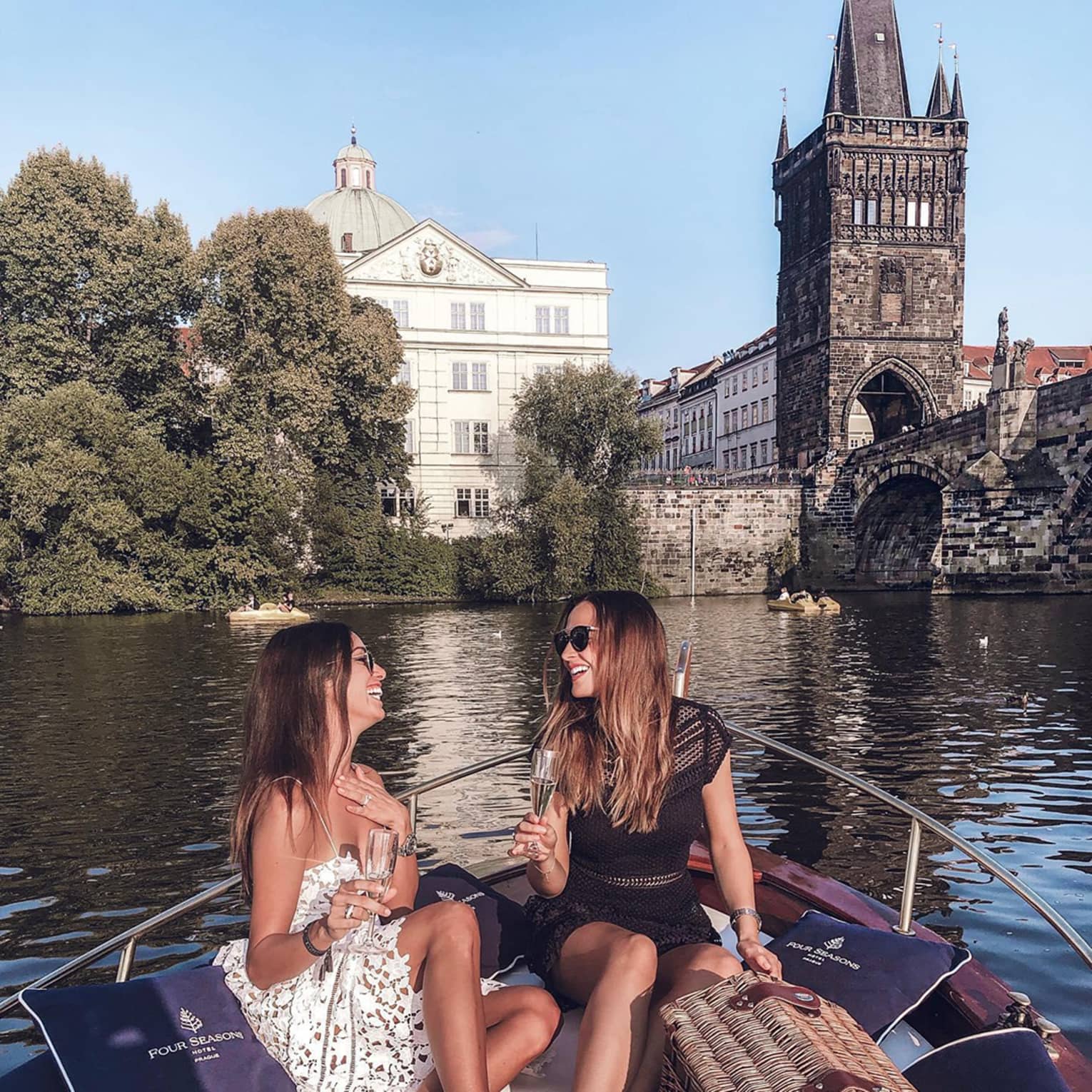 Two people sitting on a boat, enjoying champagne, with a wicker picnic basket and historic buildings and a bridge in the background.