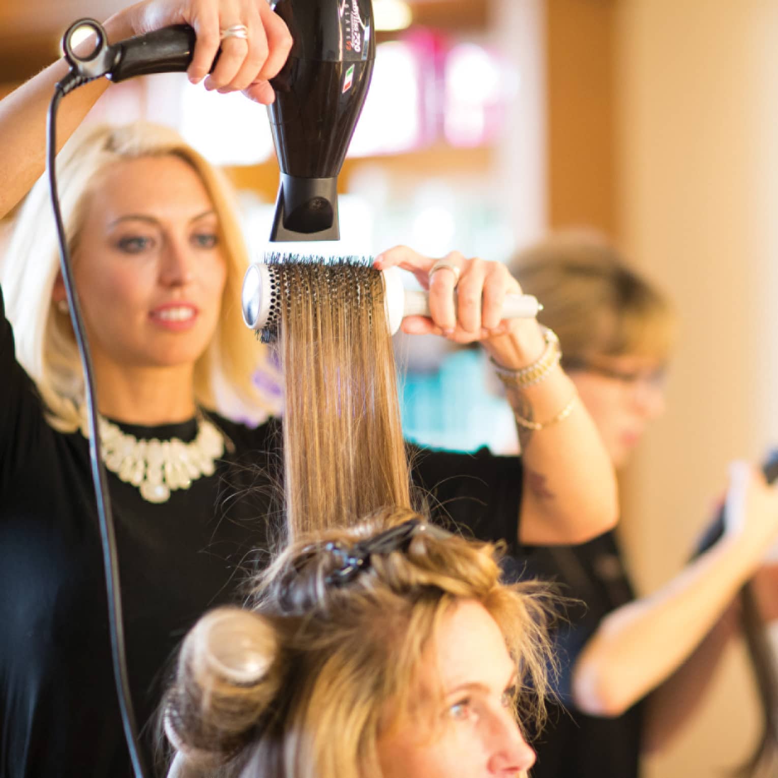Hairstylist with hairdryer pulls back woman's hair with round brush in salon
