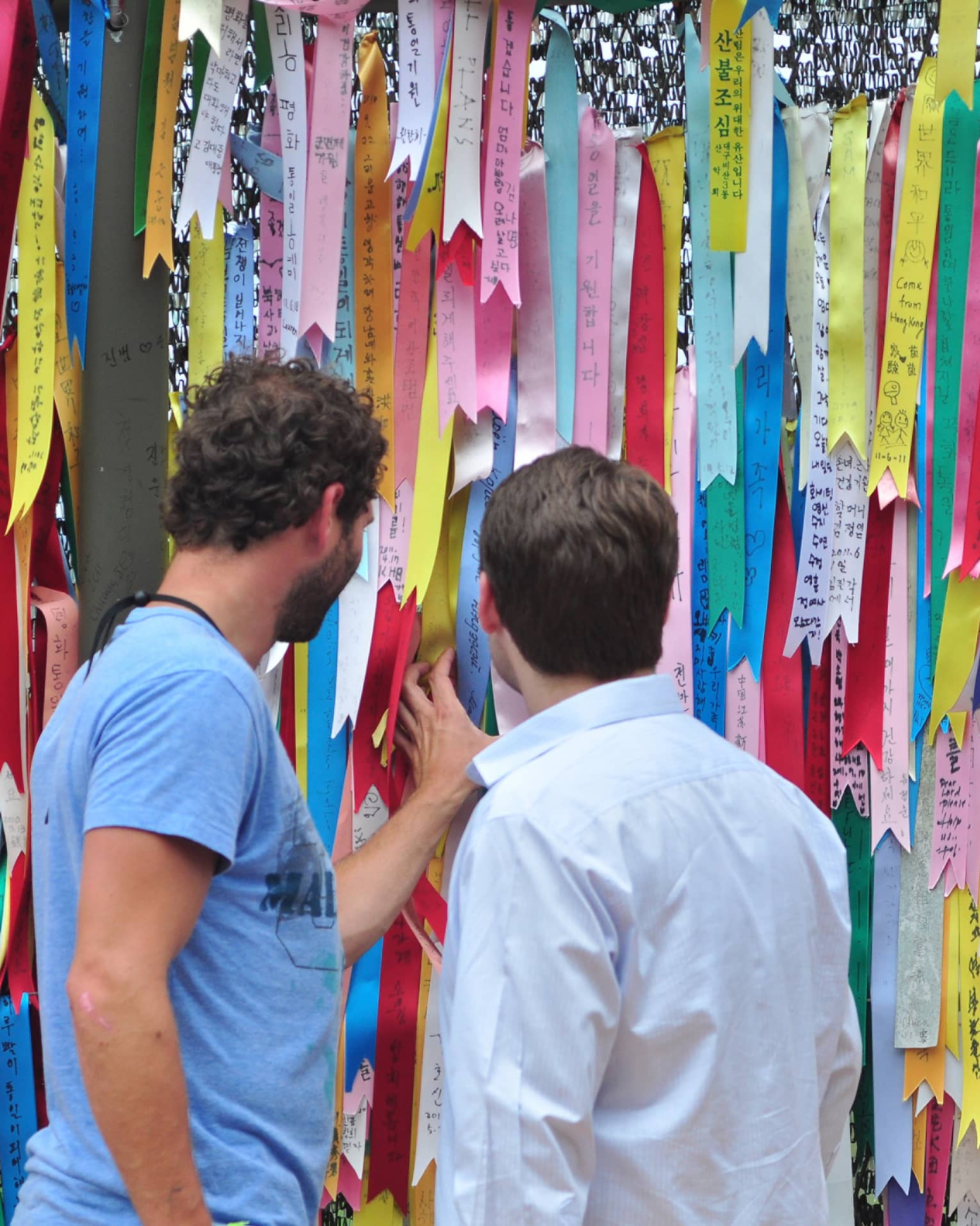 Two men looking at barbwire fence entirely covered with colourful ribbons with Korean text expressing wishes for world peace.