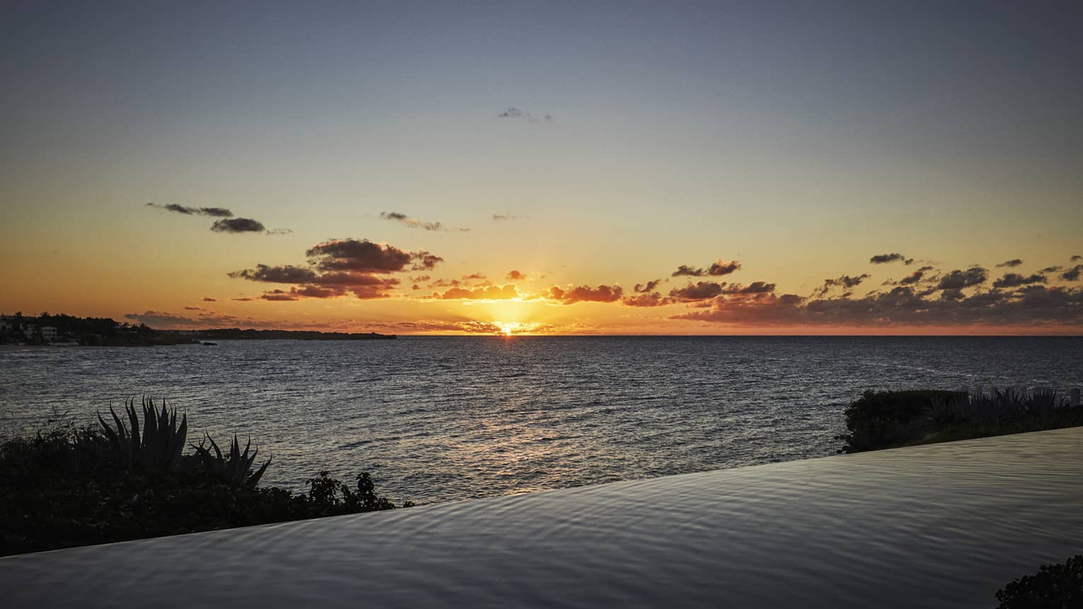Serene ocean sunset view with an infinity pool in the foreground and silhouetted coastline.
