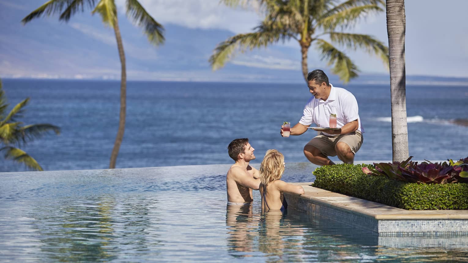 Server in white uniform with tray passes a cocktail to man and woman at edge of infinity swimming pool