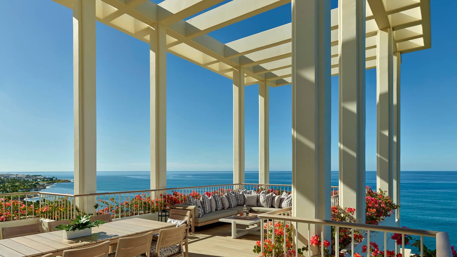 A spacious terrace with a pergola overlooking the ocean, featuring a large dining table, comfortable seating and vibrant flower arrangements under a clear blue sky