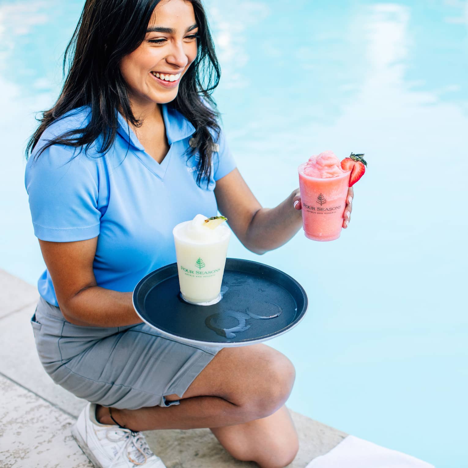 Staff member smiling in blue polo shirt kneeling at poolside with tray and two frozen drinks