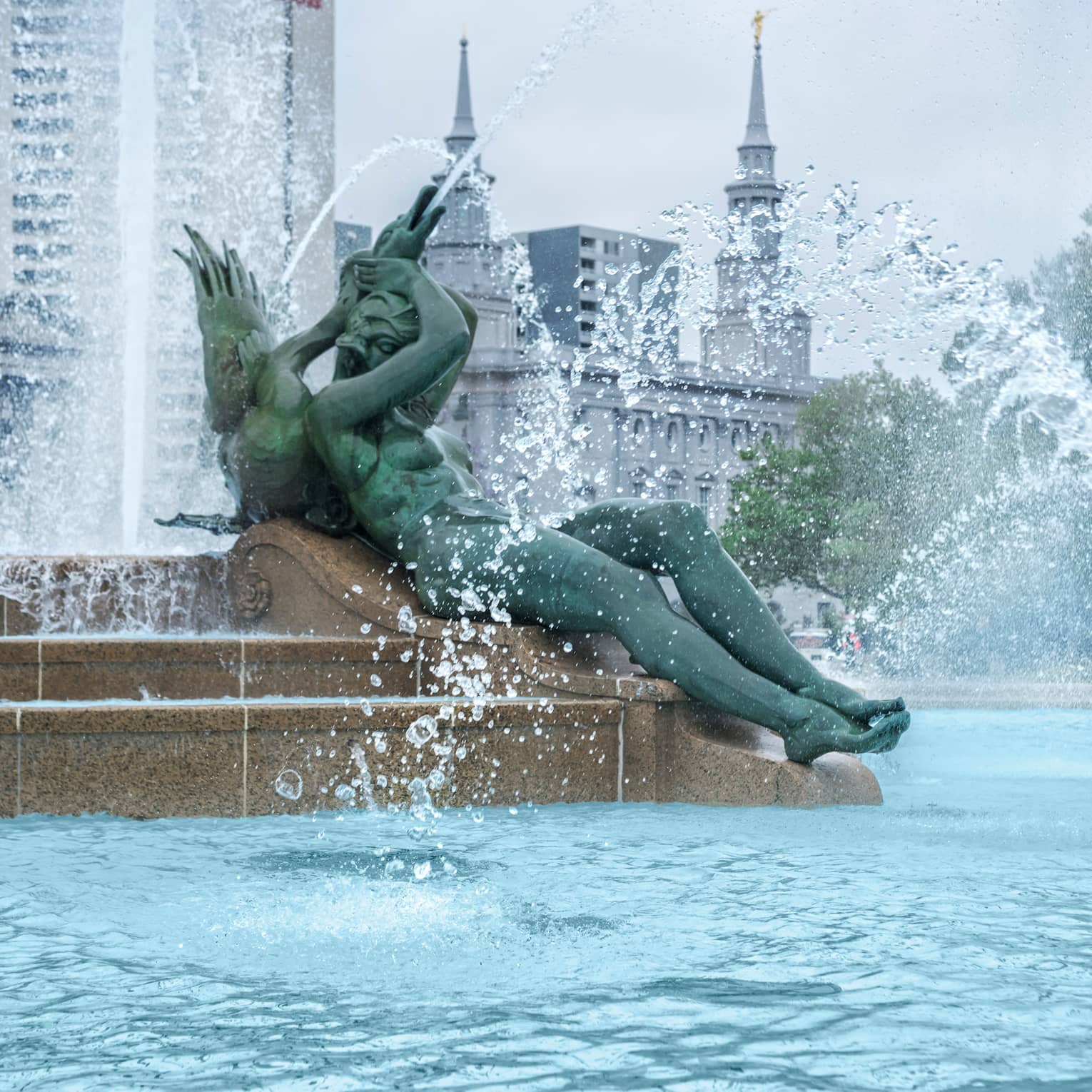 Large statue of woman leans back against centre of fountain, water sprays around