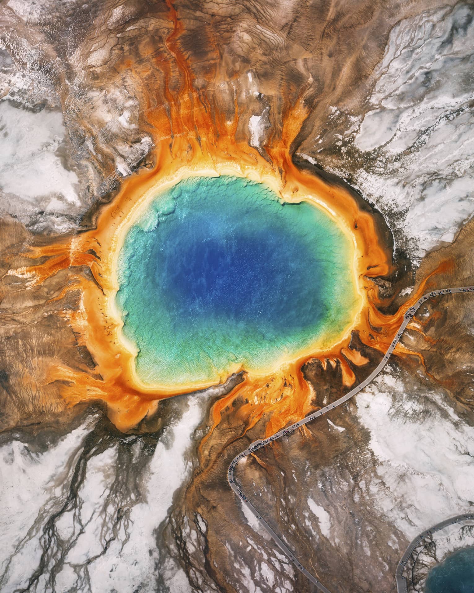 Aerial view of the Grand Prismatic Spring in Yellowstone, featuring vibrant blue, green, yellow and orange concentric rings, surrounded by rocky terrain