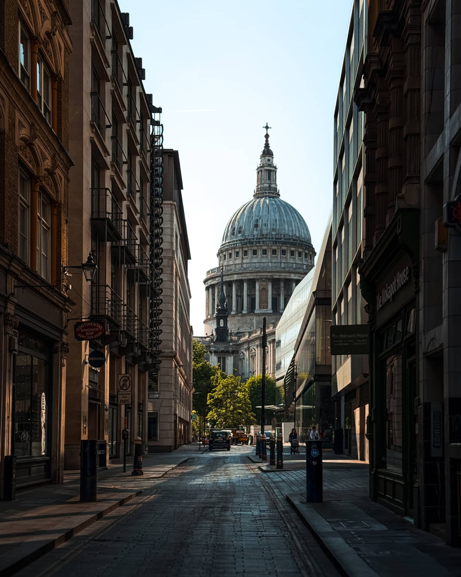 A city street in shadow, lined with buildings. The dome and spire of a cathedral rise in the background, beyond the end of the street.