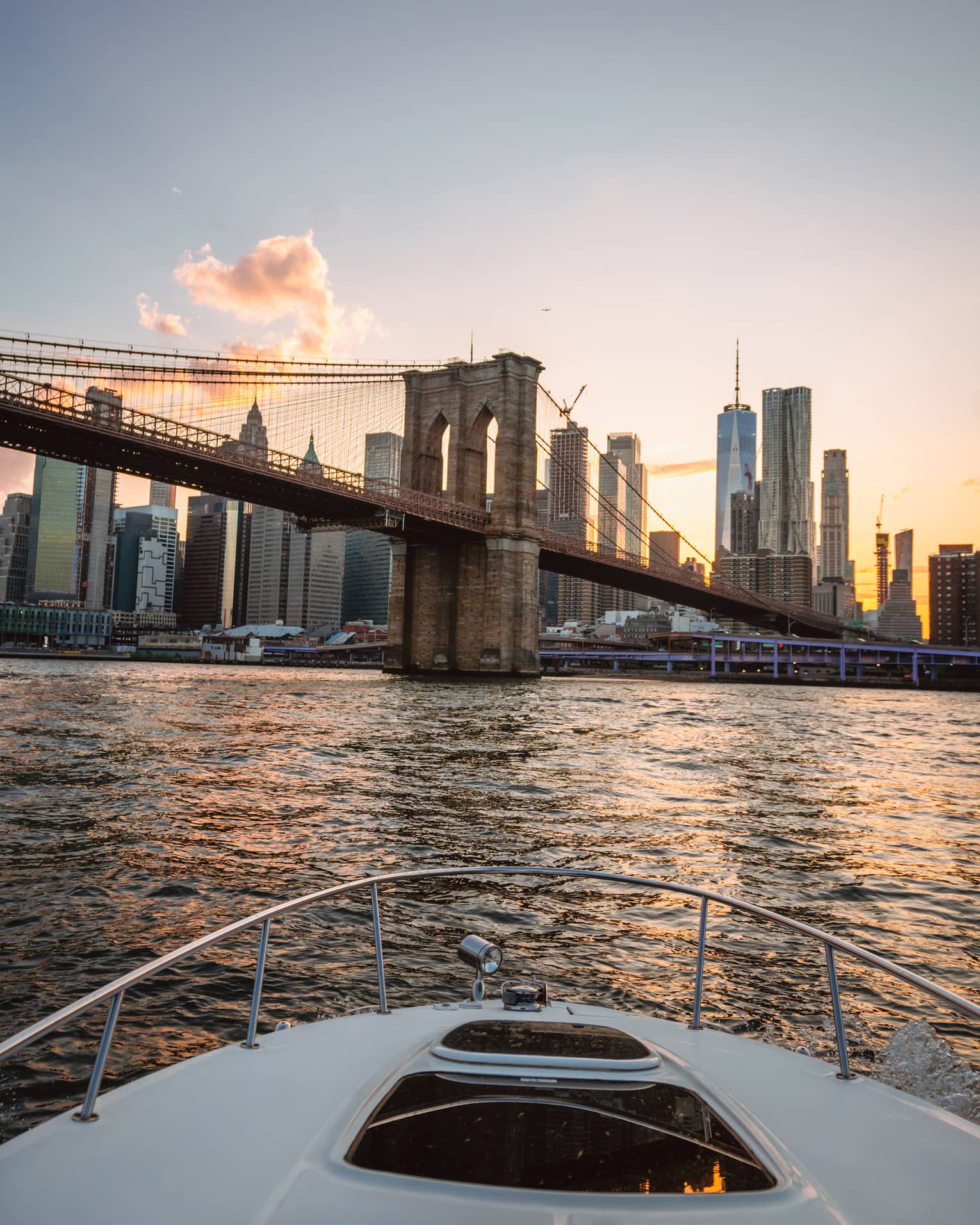 View from a boat of the Manhattan skyline behind the Brooklyn Bridge as a golden sunset reflects on the rippling water.