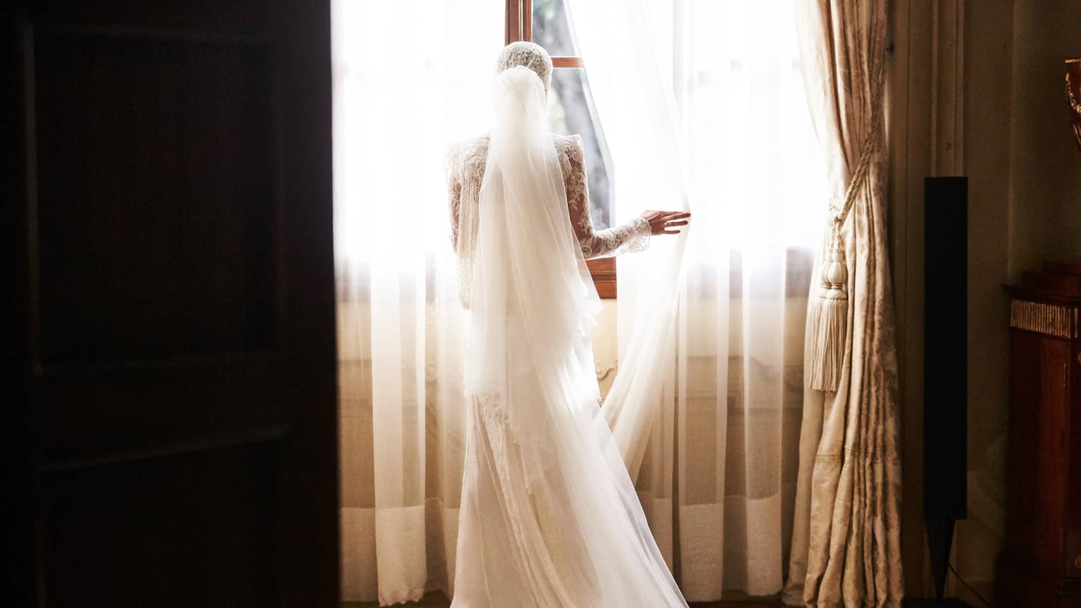 Back of bride with long white veil pulling back sheer curtain at sunny window