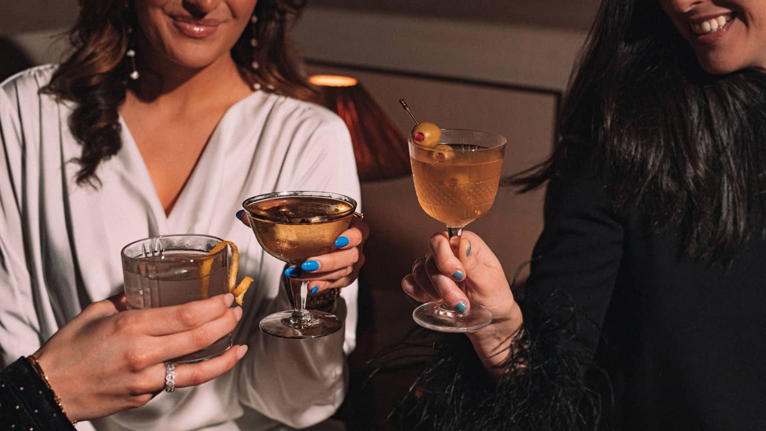 Three smiling guests share a toast with stemmed glasses and tumblers of cocktails with olive and lemon garnish.