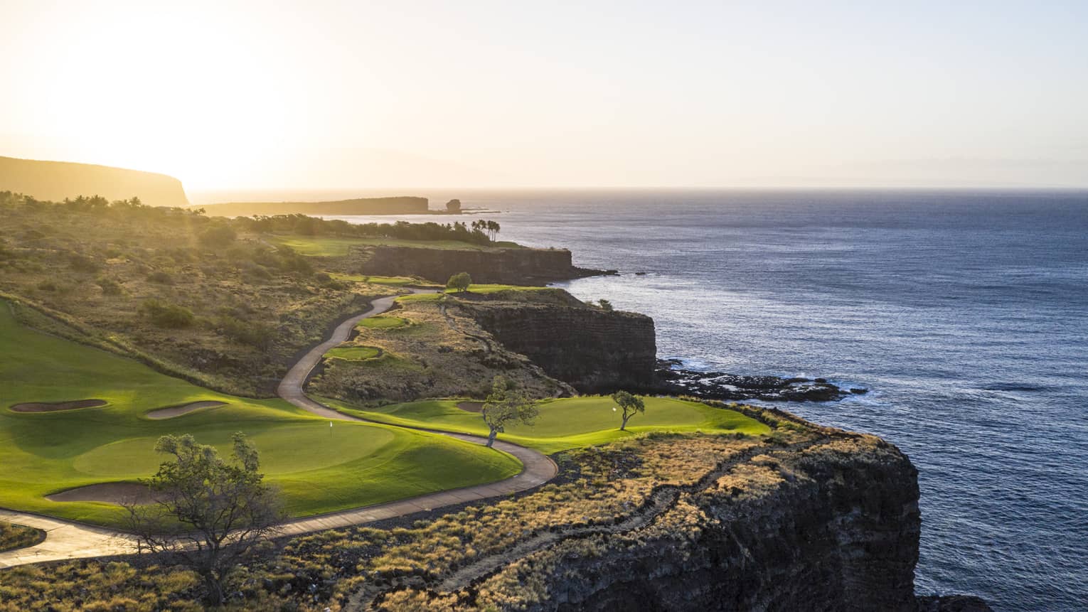 Soft sunlight filters over a pristine golf course with a winding road set on the edge of a cliff overlooking the calm ocean.