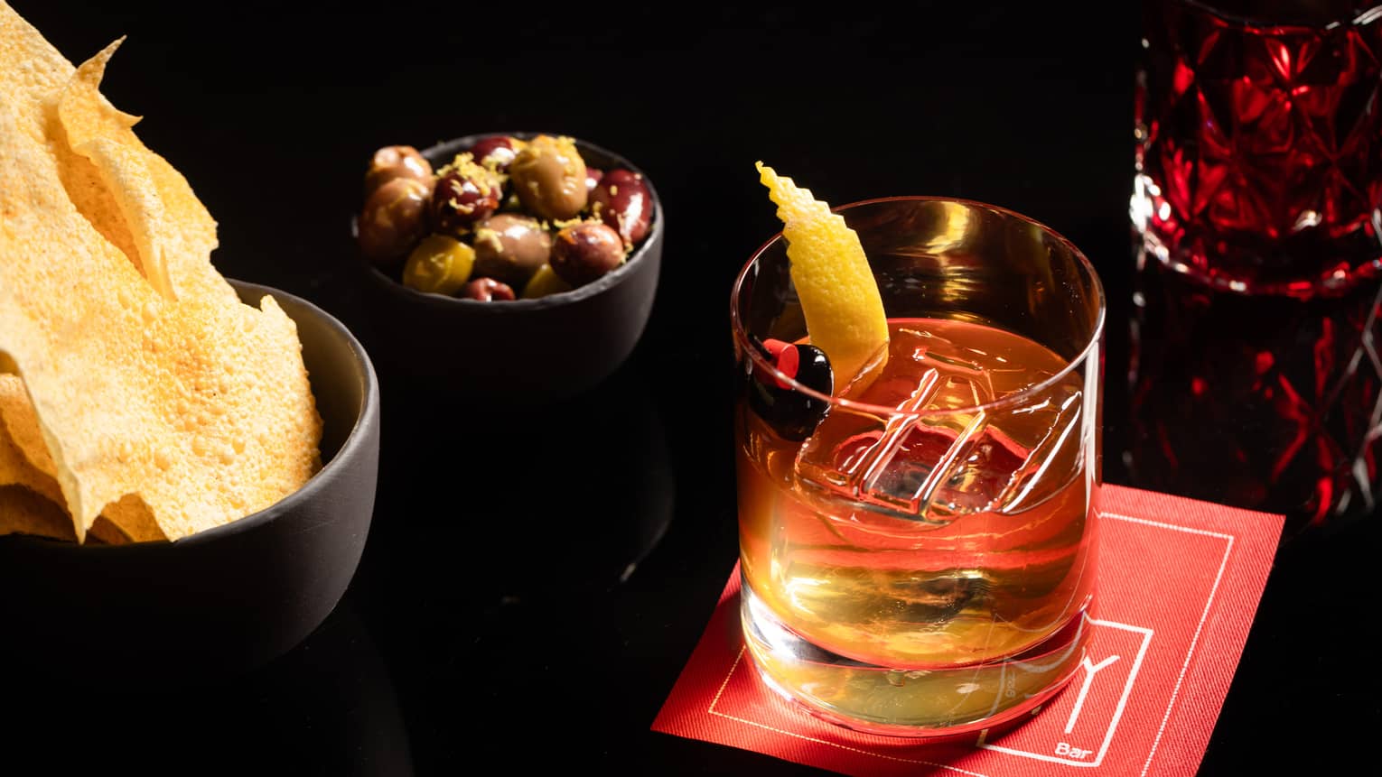 A drinking glass placed on top of a napkin with two bowls of snacks beside it.