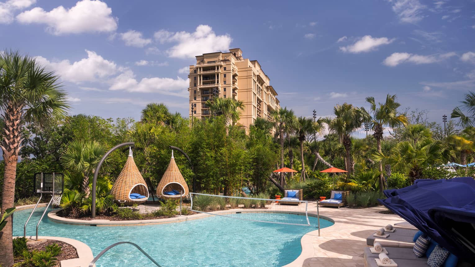 A clear blue pool featuring basketball and volleyball nets is surrounded by palm trees, covered lounges and teardrop daybeds.