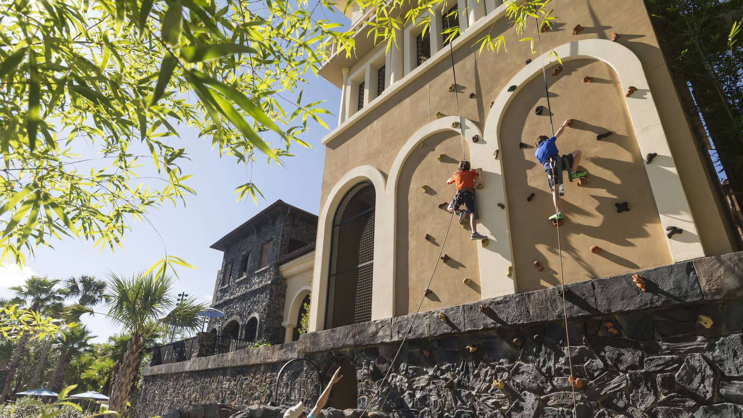 Two people climbing an outdoor rock climbing wall set along the side of a building with an instructor standing on the ground