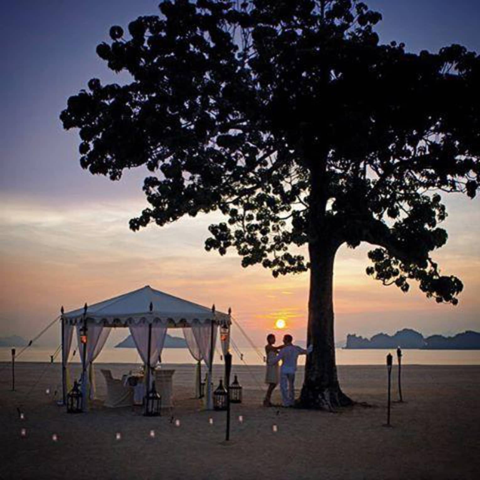 A tent, dining table and lanterns are set up oceanside under a large tree while a couple stands nearby watching the sunset
