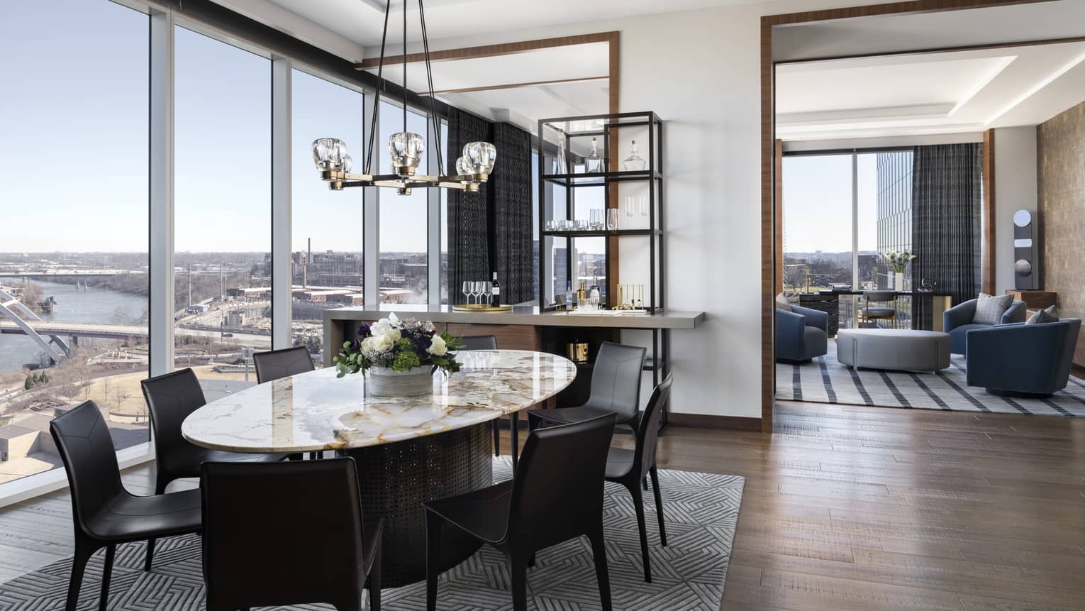 Spacious dining area with a marble table, leather chairs and a chandelier, adjacent to a living room with blue chairs and a river view through floor-to-ceiling windows