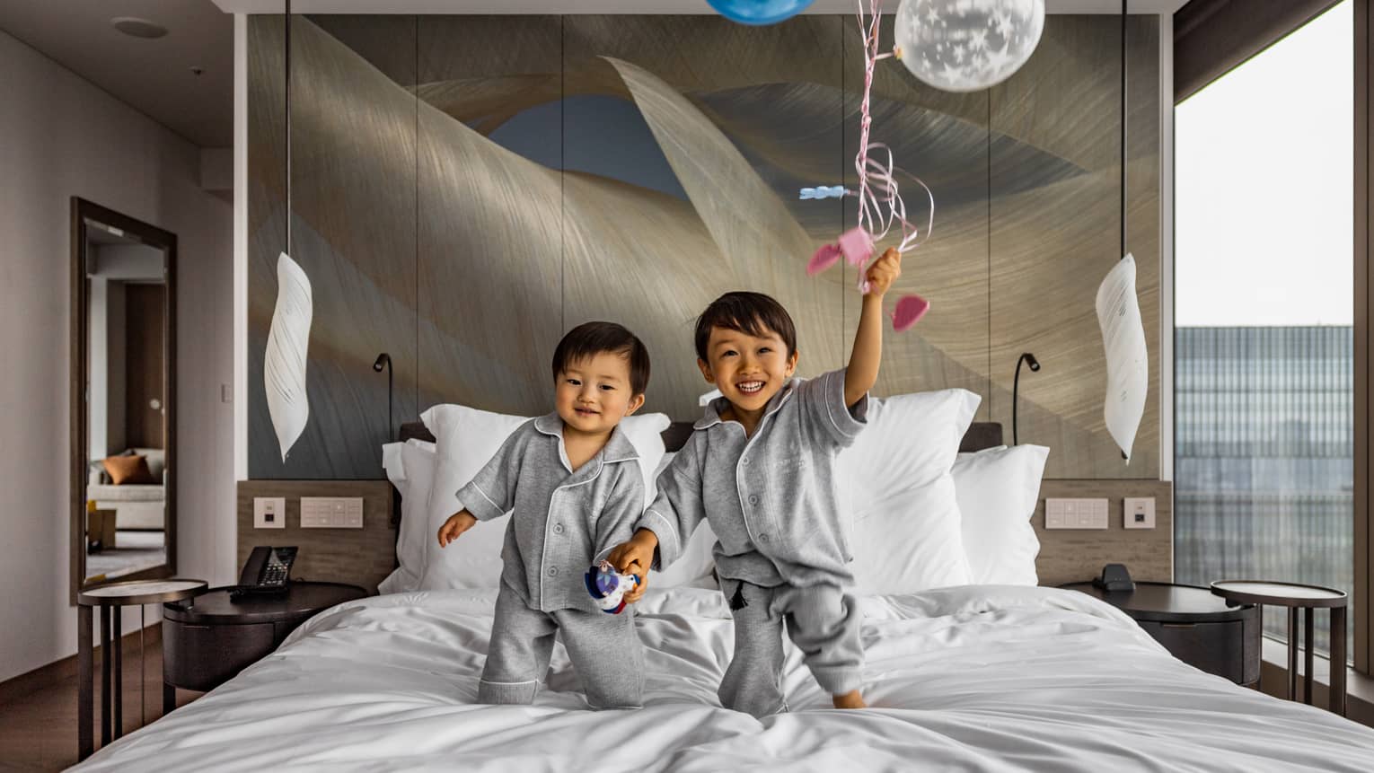 Two young children in grey pyjamas hold hands while standing on a bed in a bright room; one smiles and holds a clear balloon.