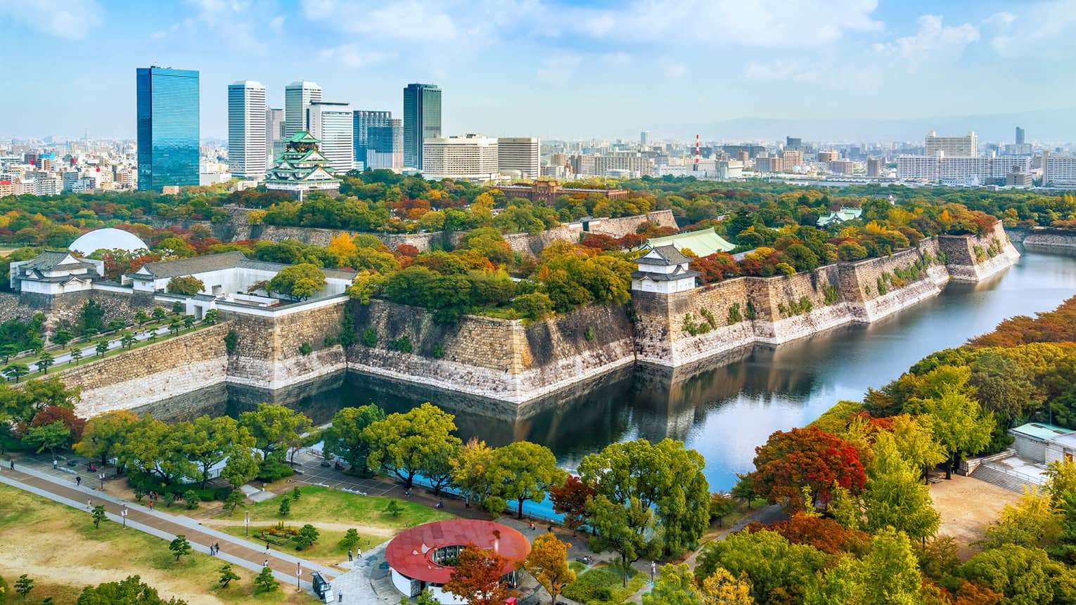 Osaka Castle rises amid lush autumn-coloured trees within a stone fortress surrounded by a moat and modern high-rises.