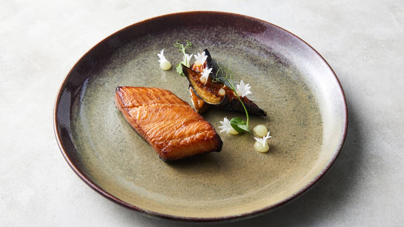 Black cod plated with a floral garnish on a brown stoneware plate rimmed in burgundy