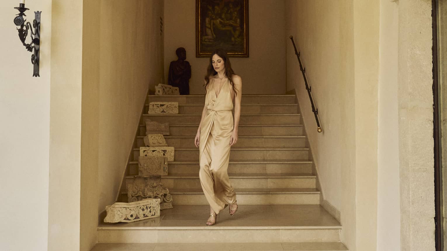 Woman wearing Champagne-coloured satin dress walks down the stairs in an arched walkway