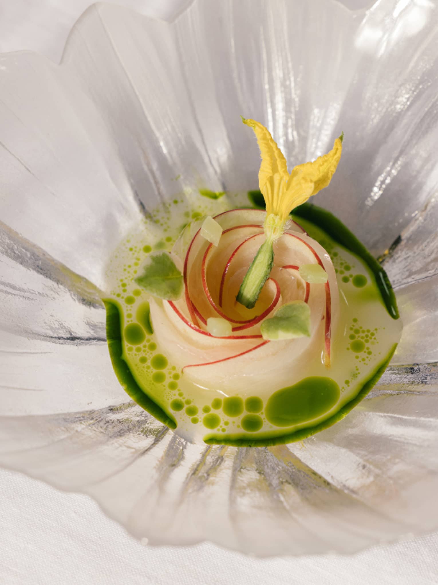 Elegant dish arranged as a rose, topped with a yellow edible flower, in a green herb-infused broth