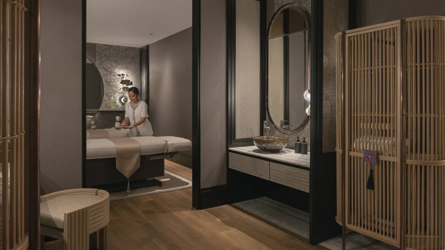 A tray is gently placed on one of two massage tables in a warmly lit spa room with soft grey walls and natural wood décor