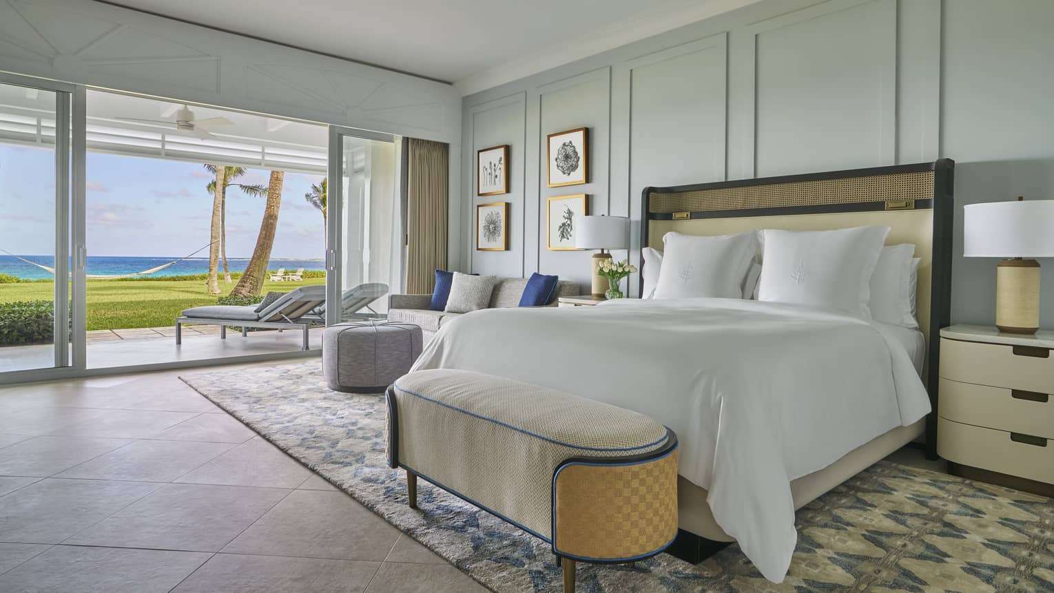 Luxury oceanfront bedroom with a large bed, cozy seating and sliding glass doors opening to a private patio with lounge chairs