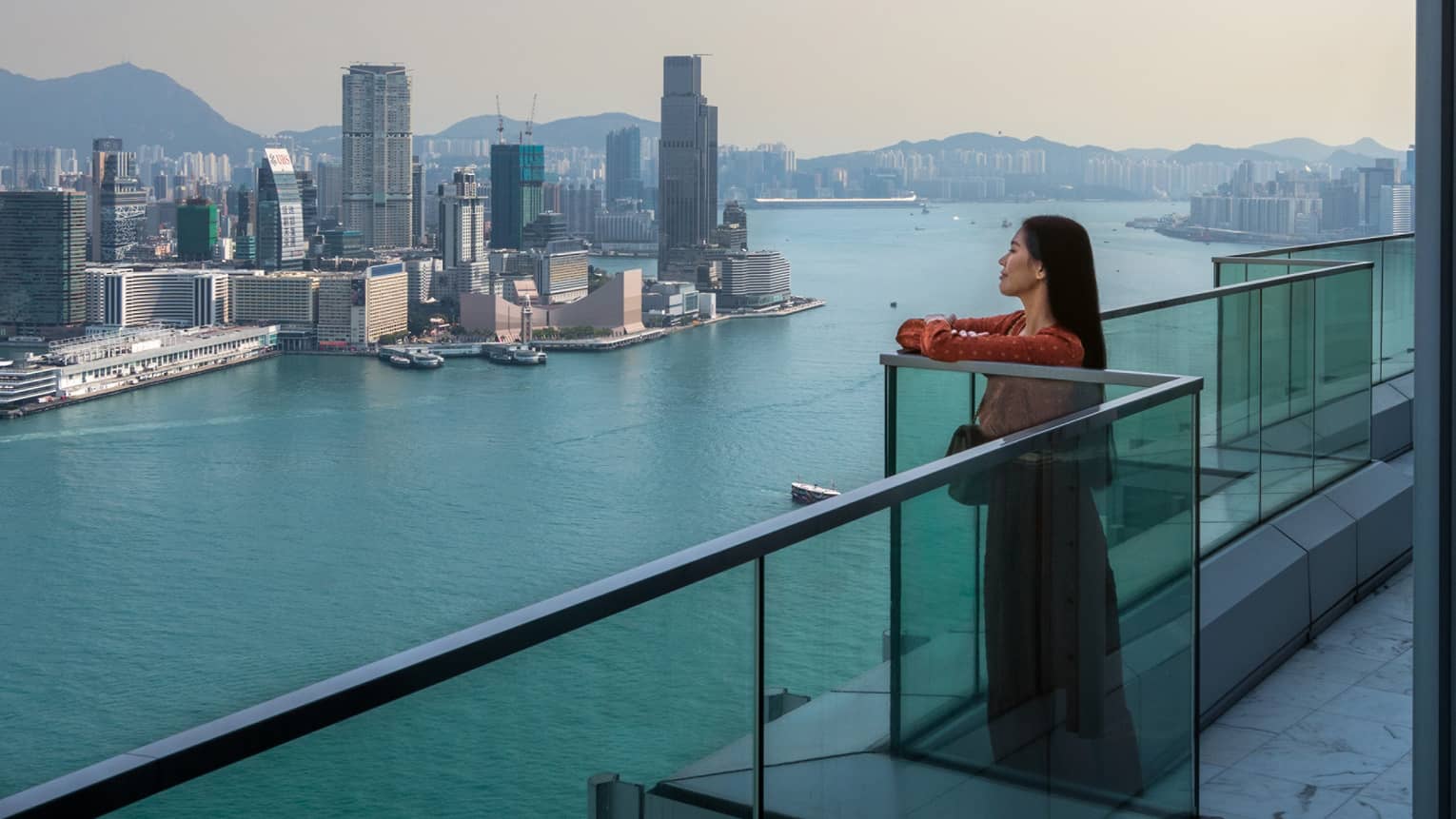 A woman standing on a balcony looking out at water and a city.
