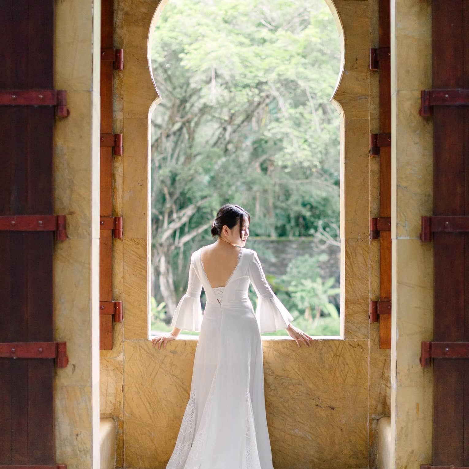 ,A woman in a white wedding dress stands in front of a large window overlooking green foliage