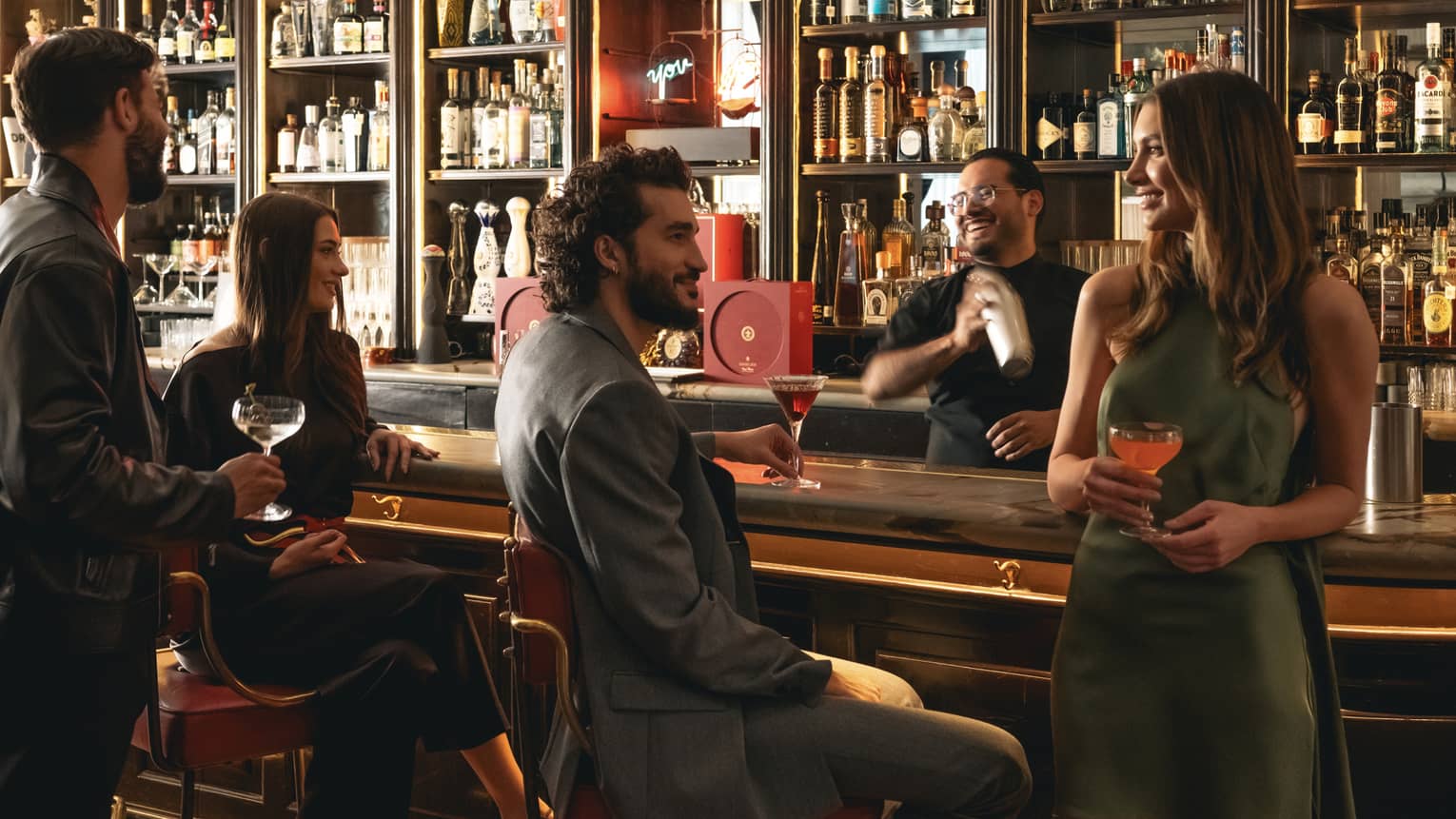People socialize at a bar counter; a bartender making a drink in a shaker is in the background.