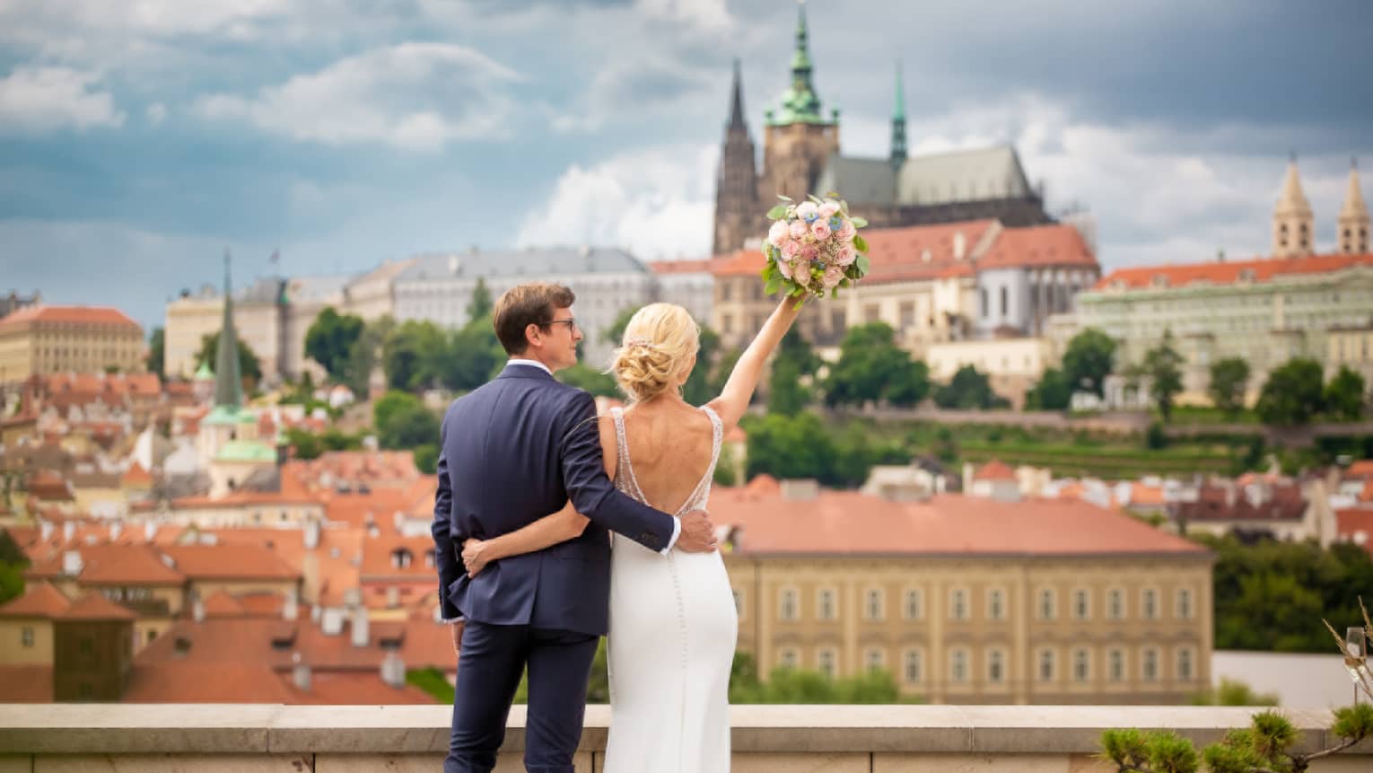 Back of groom and bride lifting bouquet of pink roses towards Prague city view