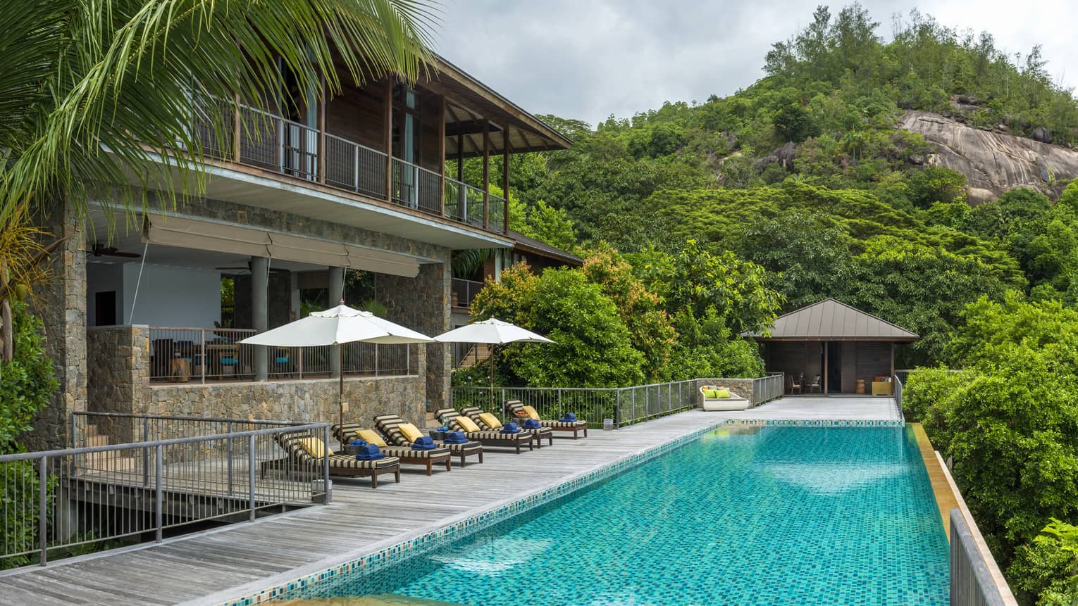 Row of patio chairs and umbrellas on balcony beside long blue swimming pool, mountain in background
