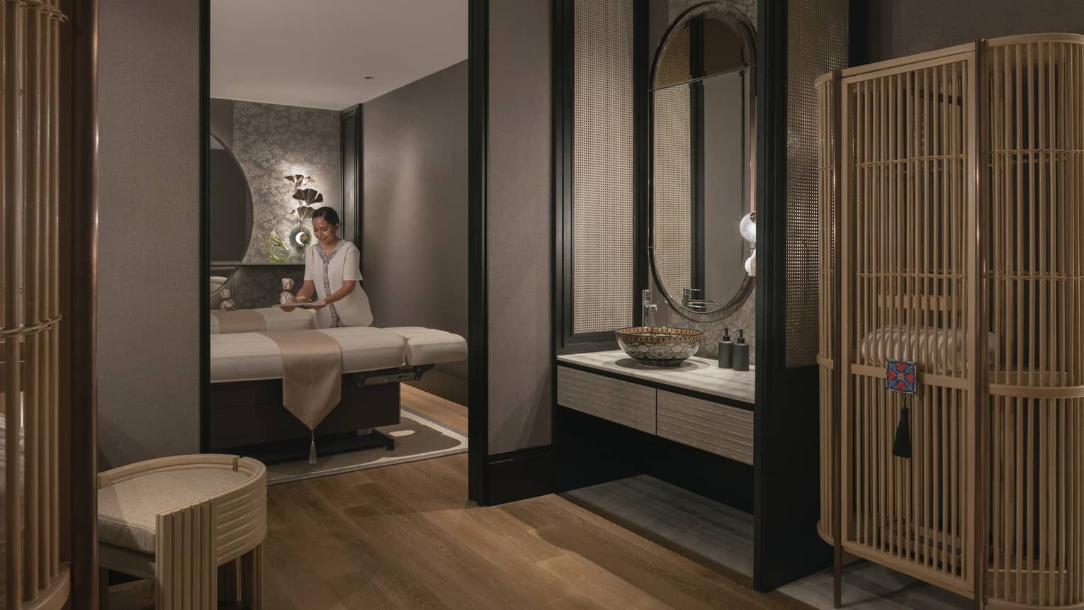 A tray is gently placed on one of two massage tables in a warmly lit spa room with soft grey walls and natural wood décor