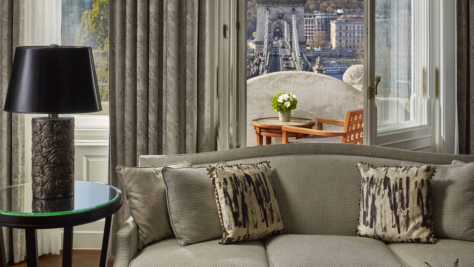 Sofa with patterned pillows facing a balcony with table and chairs overlooking a historic bridge