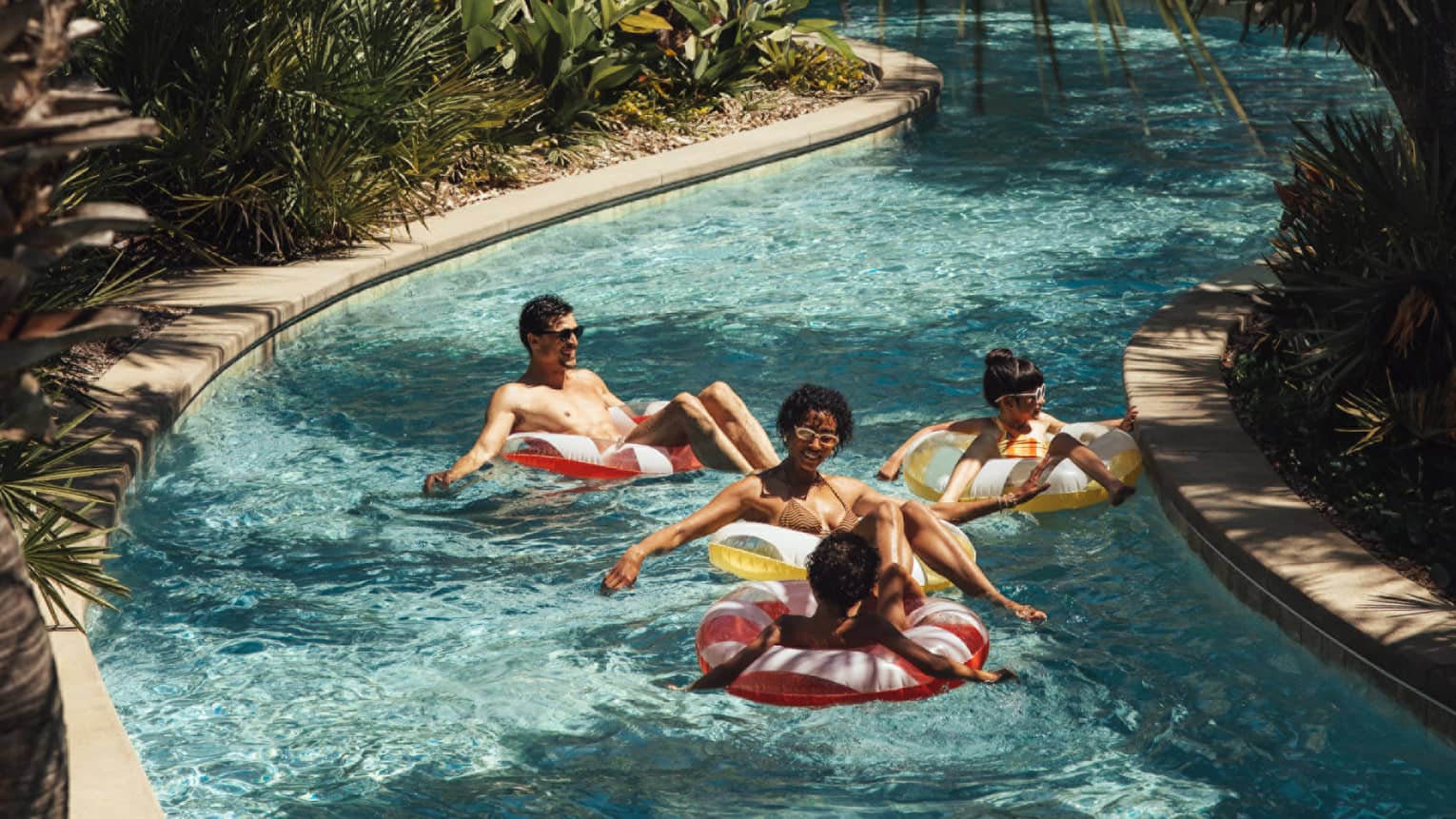 Two adults and two young children float in inner tubes down a lazy river surrounded by palm trees