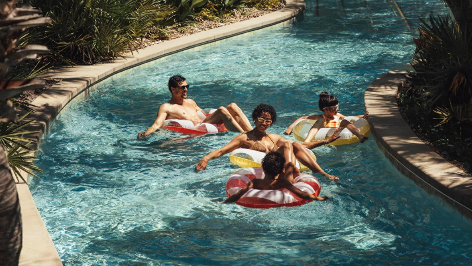 Two adults and two young children float in inner tubes down a lazy river surrounded by palm trees