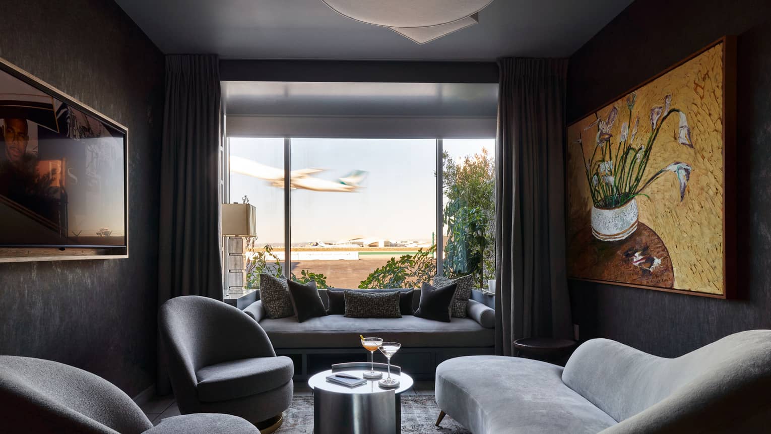 A cozy, contemporary interior space decorated in shades of grey, with a large window, plush furniture and art on the walls