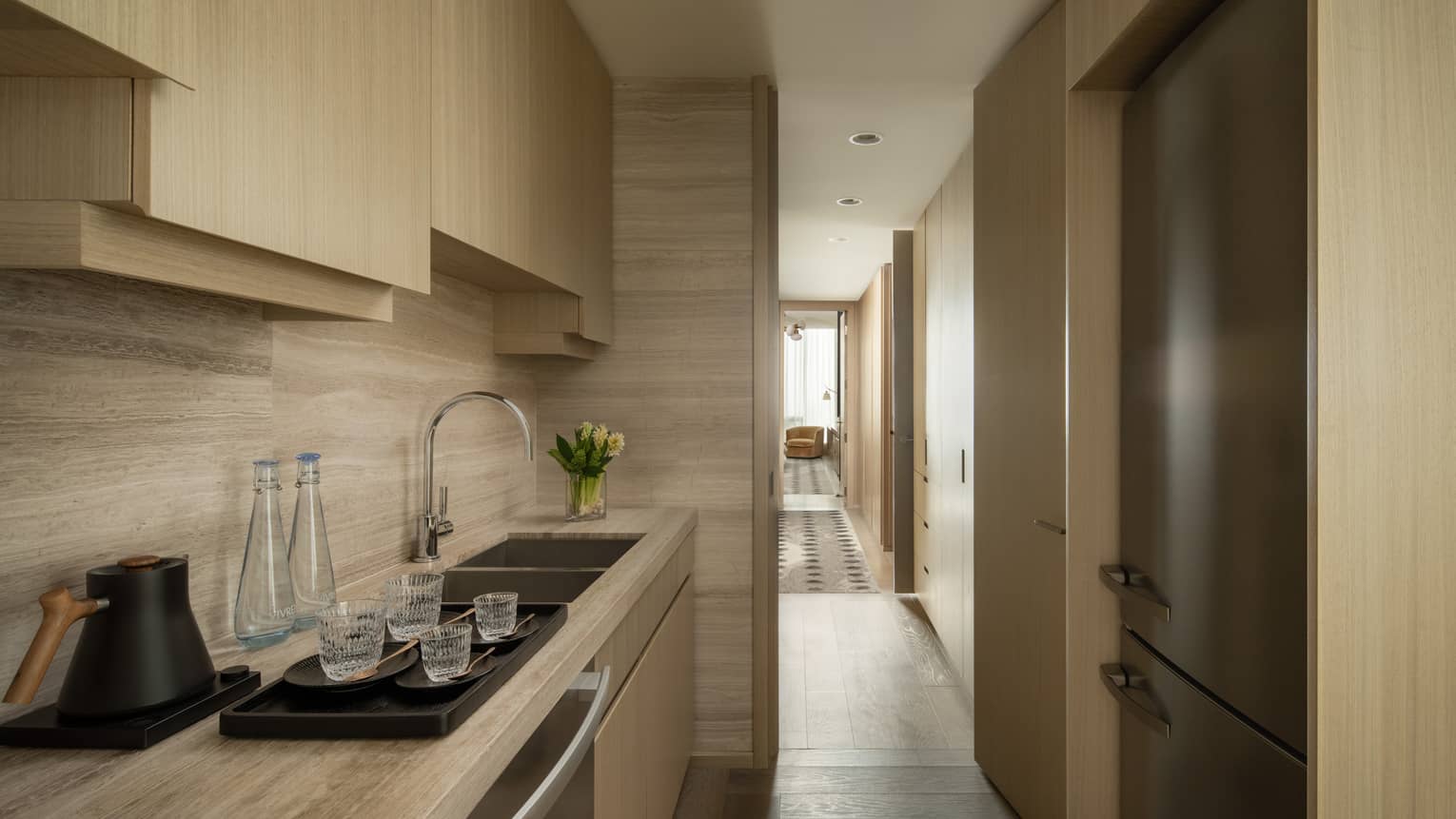 Kitchen area of a hotel suite with beige wood cabinets, refrigerator, sink, black kettle, water bottles and a tray with glasses