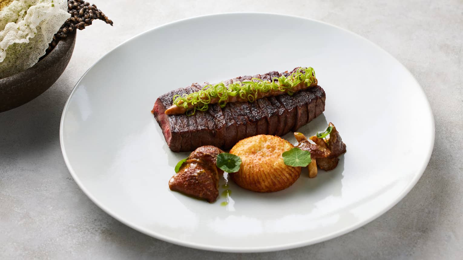 Grilled Wagyu beef strip-loin topped with a green garnish and plated next to cooked mushrooms on a white dish with
