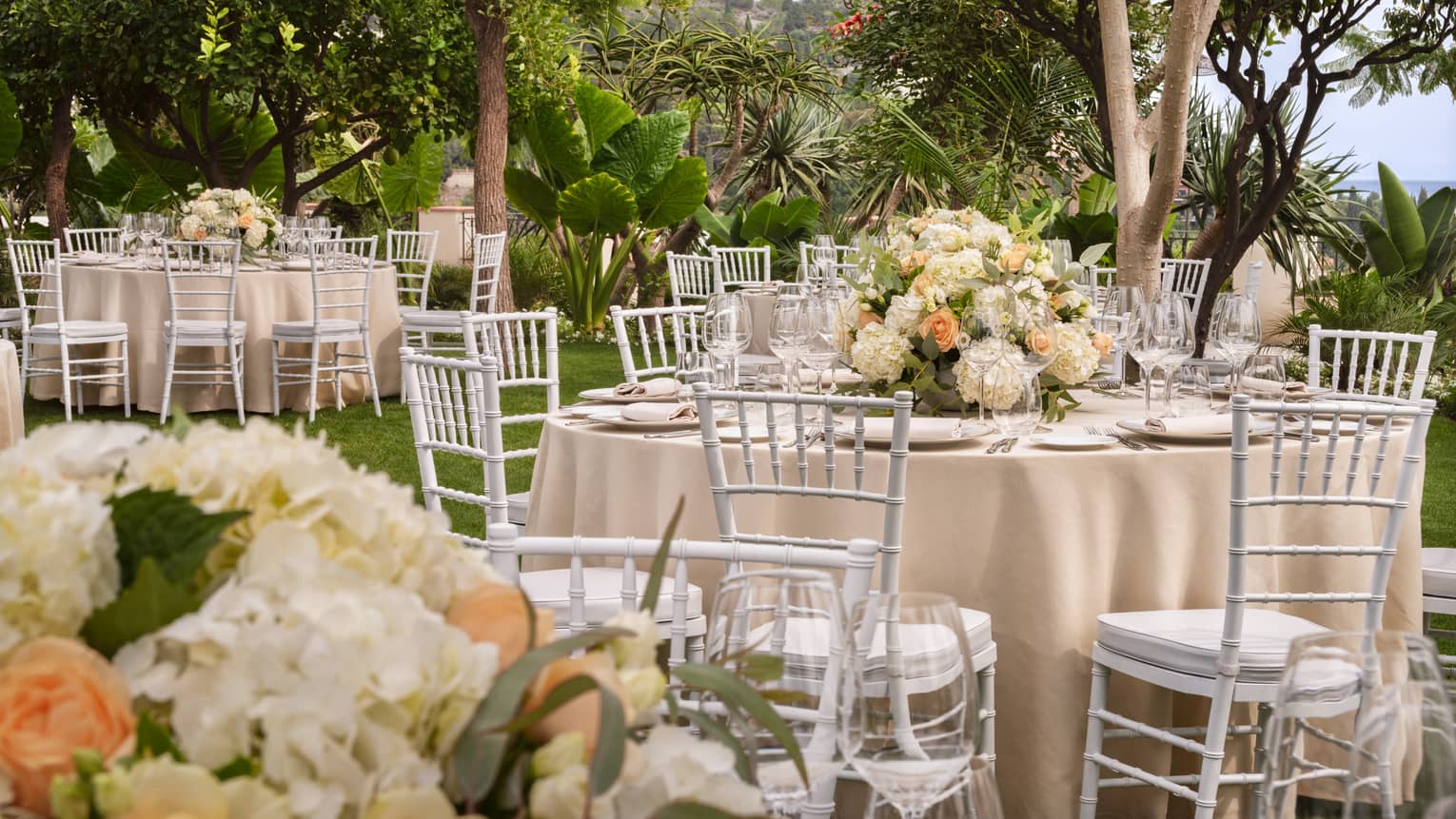 Outdoor wedding reception featuring white chairs and tables adorned with peach and ivory flowers in a garden setting