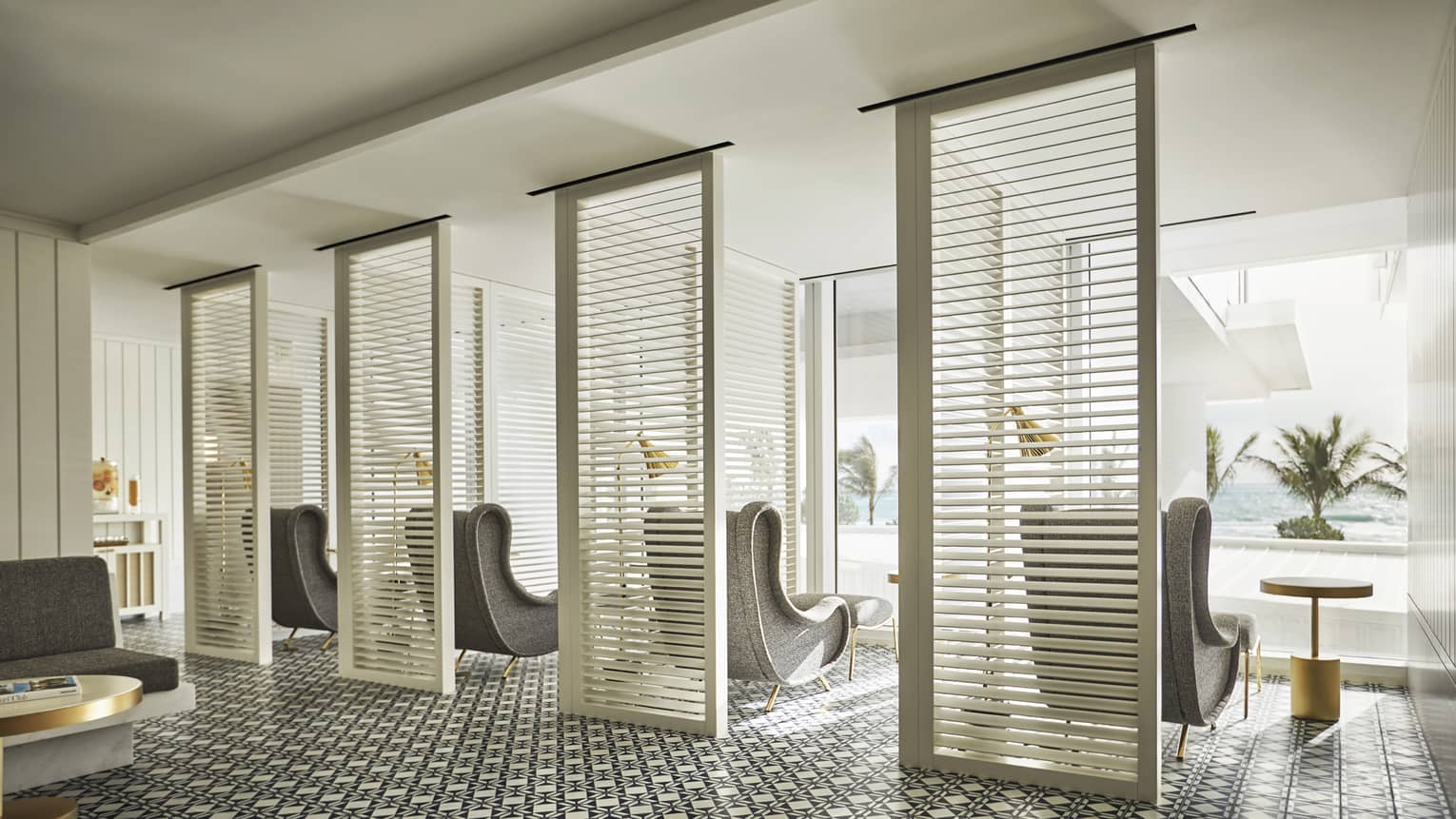 Luxury spa area with patterned tile flooring, partitioned seating space, gray armchairs and floor-to-ceiling windows overlooking ocean and palm trees
