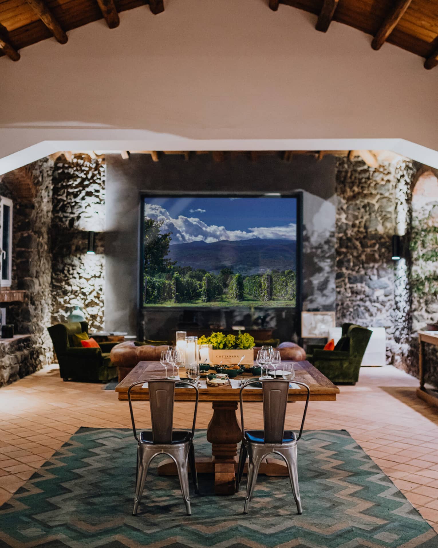 A wooden dining table set with floral arrangements and glassware, in a room with a window overlooking mountains and a vineyard