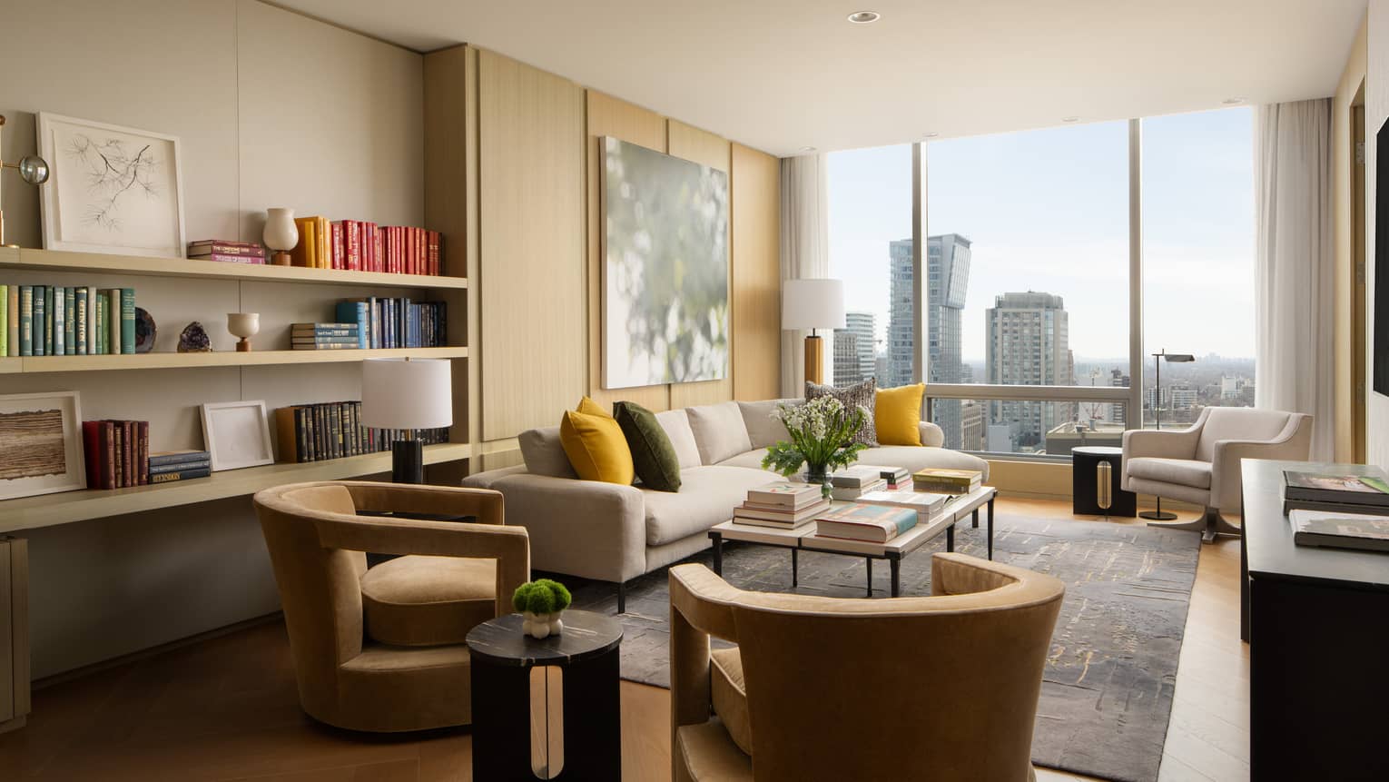 Luxury suite lounge with armchairs, cream sofa, mustard and green pillows, built-in bookshelves and floor-to-ceiling windows offering a city skyline view