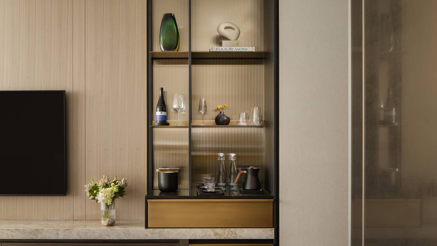 Stylish minibar with glass shelves displaying wine, glasses, and decorative items, next to a TV and vase with fresh flowers