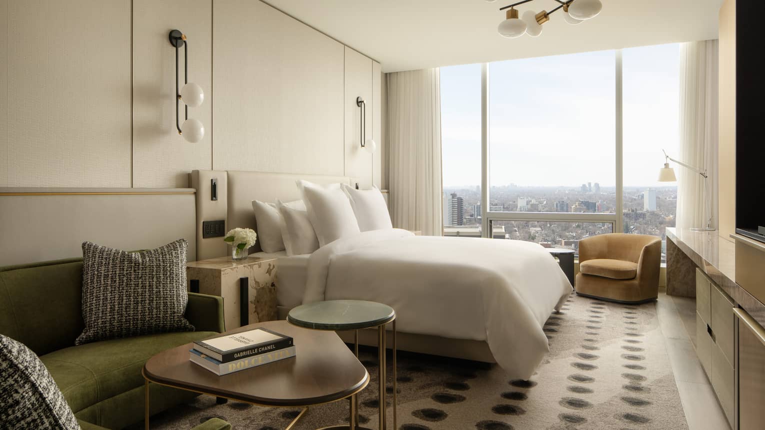 Suite bedroom with a king-size bed, green sofa, side and coffee table, armchair and floor-to-ceiling windows with city views