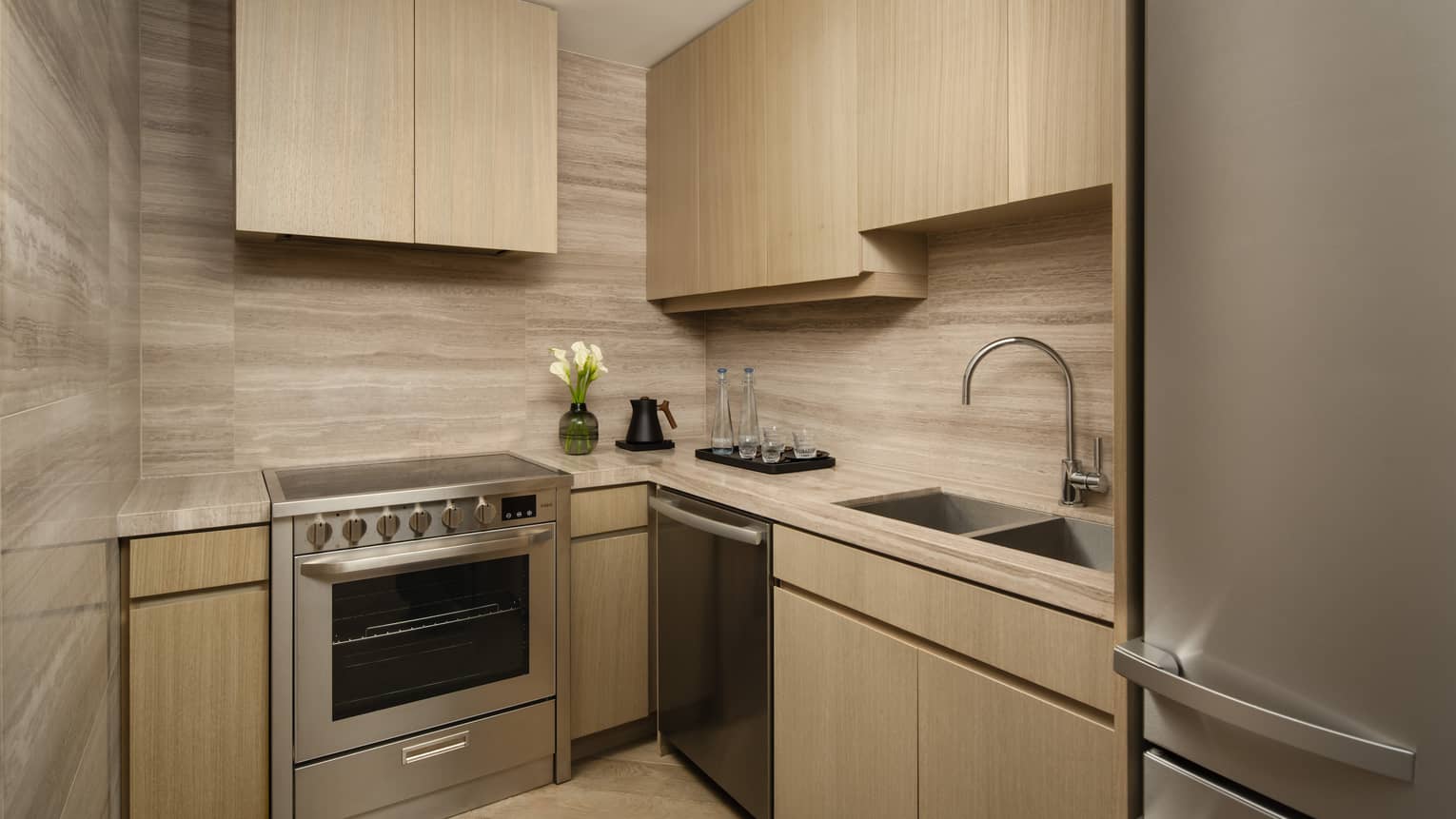 Compact kitchen with light wood cabinetry, sink, dishwasher, stove, and fridge, featuring a small vase and water glasses on the counter