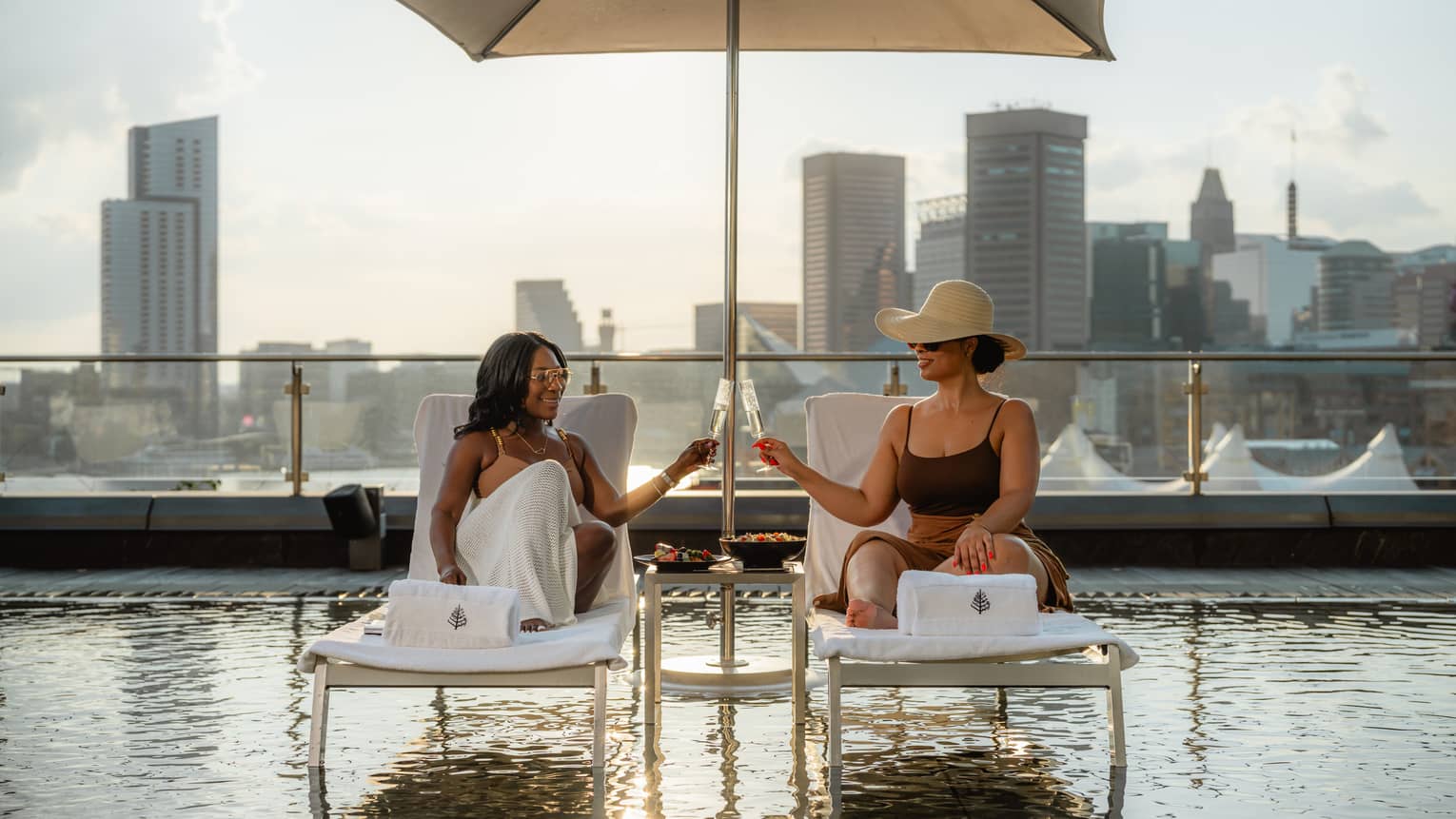 Two guests have a toast with drinks in stemmed glasses, while they sit on lounge chairs under a sun umbrella over a rooftop pool, with city skyline views in the background.