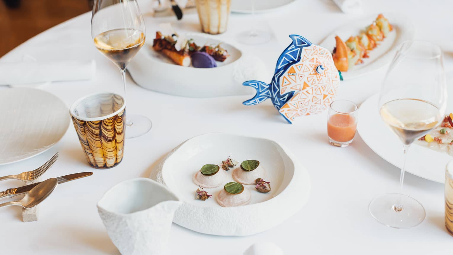 A white round table with a blue and orange centerpiece is set with plates, utensils, napkins, tumblers and clear stemmed glasses. Serving plates hold colourful food items.